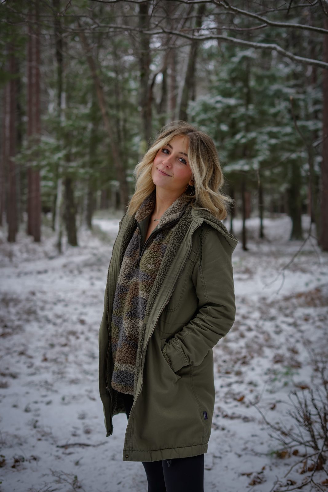 Ashlyn smiling in an olive parka in a snowy pine forest